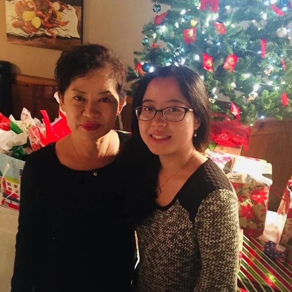 Two women pose together beside a brightly lit Christmas tree surrounded by wrapped presents in a cozy home.