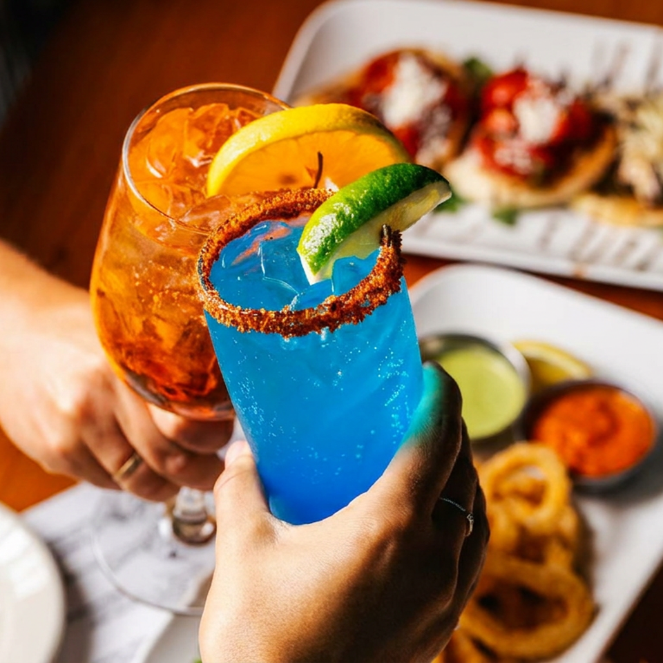 Two hands clinking vibrant cocktails, a blue margarita and an orange spritz, over a table of appetizers.