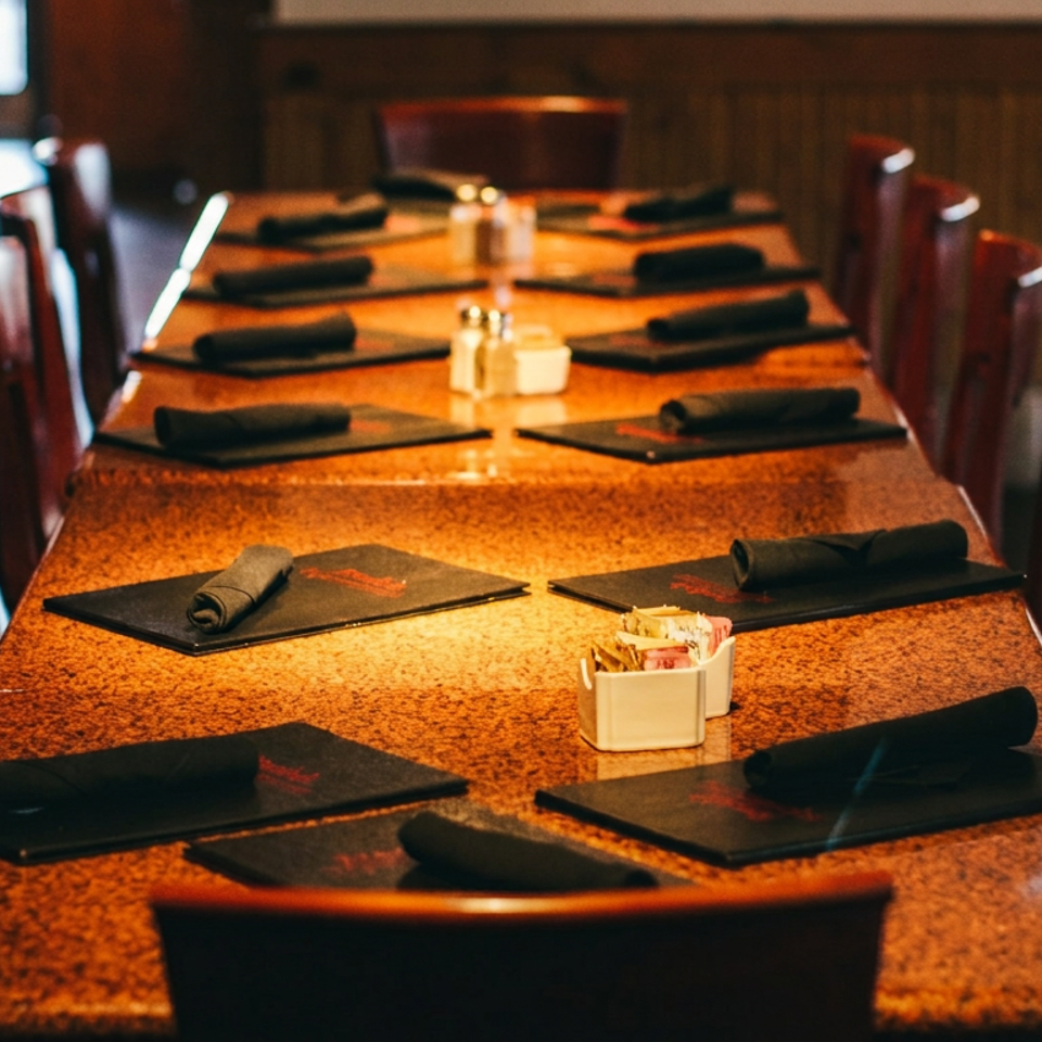 Long dining table set with black placemats, rolled napkins, salt and pepper shakers, and sugar packets.