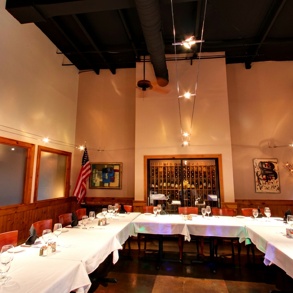 Well-lit private dining room with U-shaped table, white tablecloths, red chairs, and a wine display.