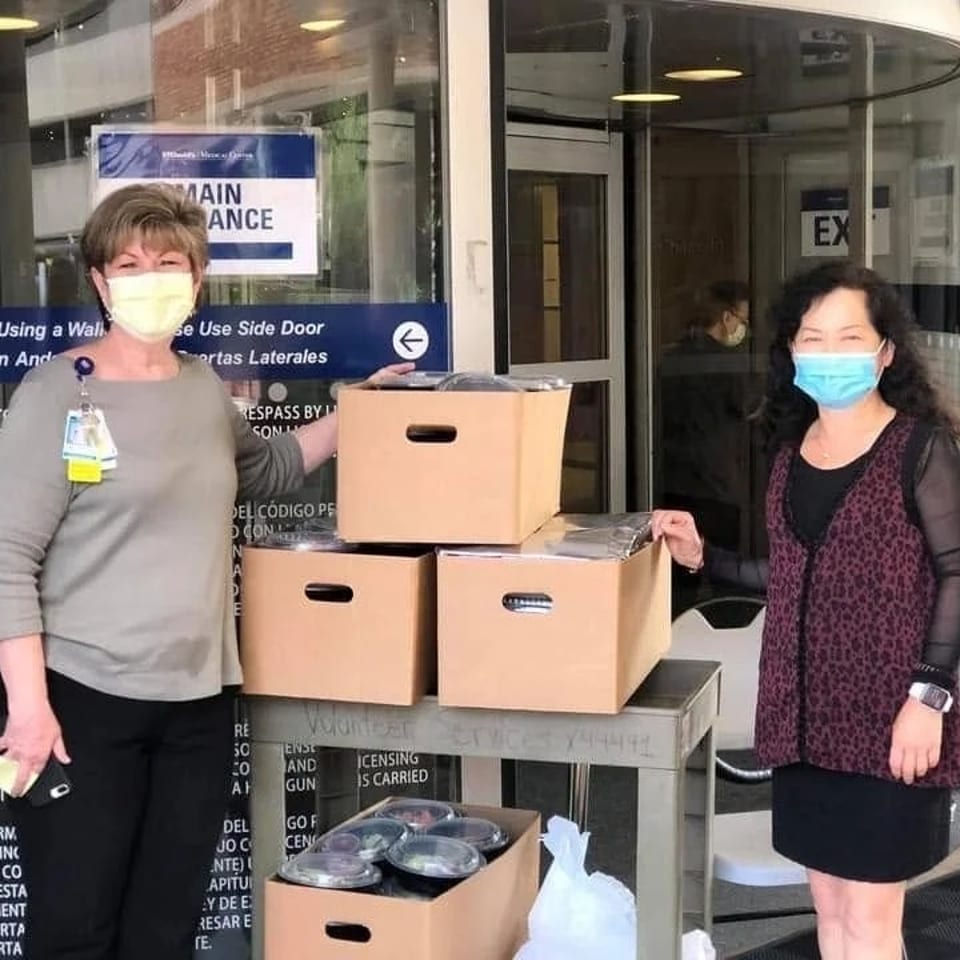 Two masked women stand beside a cart loaded with boxed meals outside a building entrance.