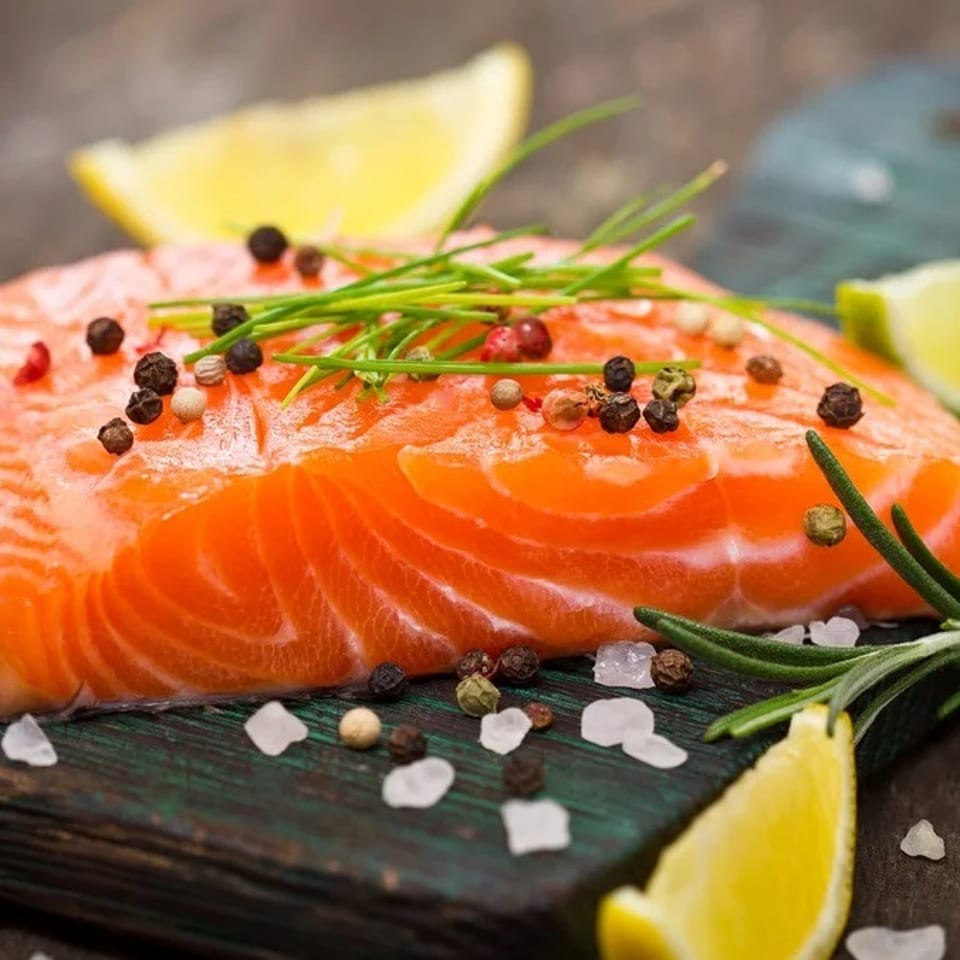 Raw salmon fillets garnished with peppercorns, chives, rosemary, coarse salt, and lemon wedges on a dark serving board.